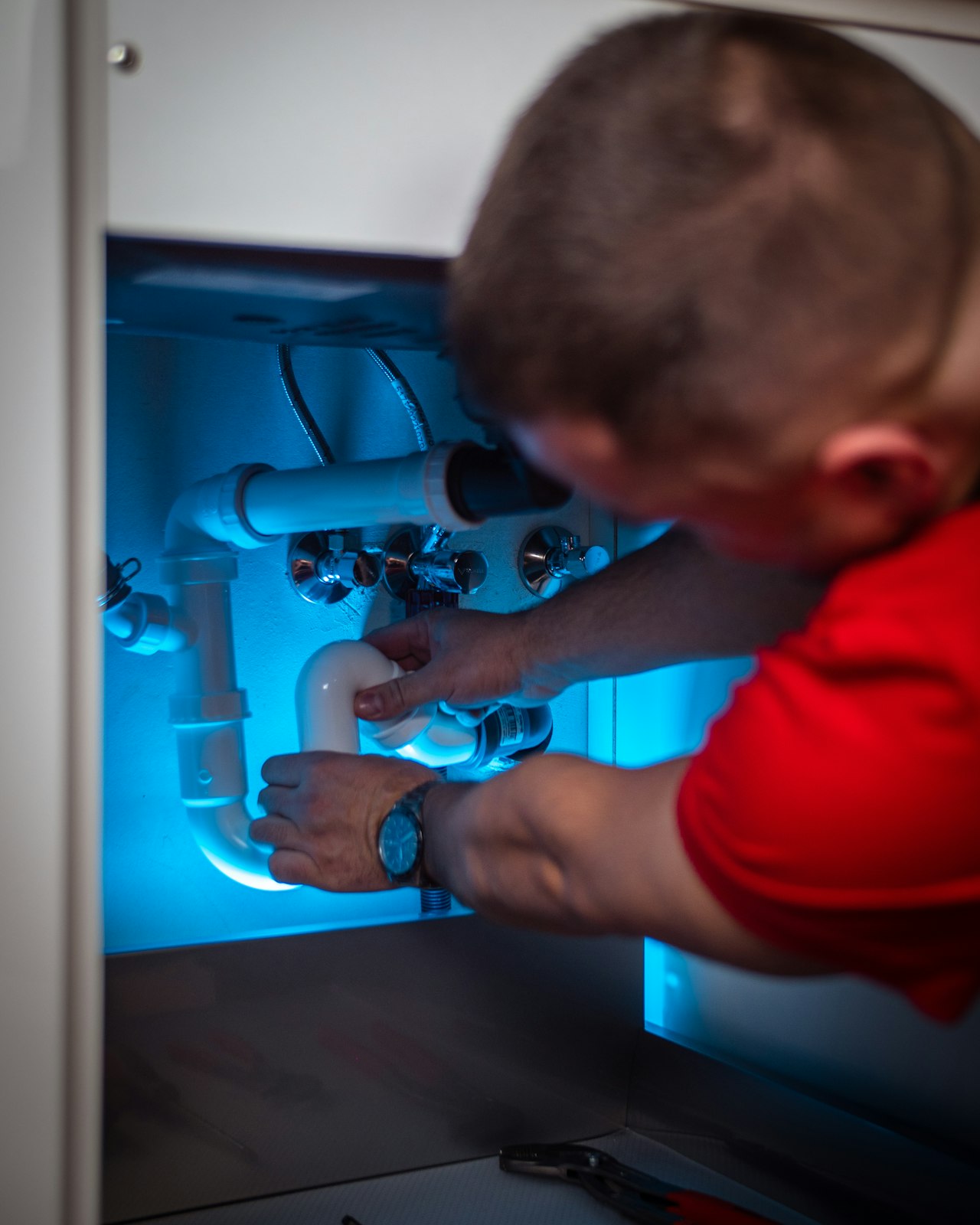 A plumber reaching into a sink cabinet to work on chrome drain fittings under bright, uncluttered daylight.